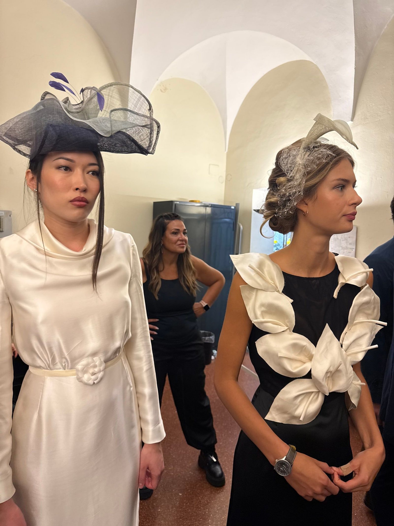 Due modelle nel backstage con copricapi e un abito nero con petali avorio